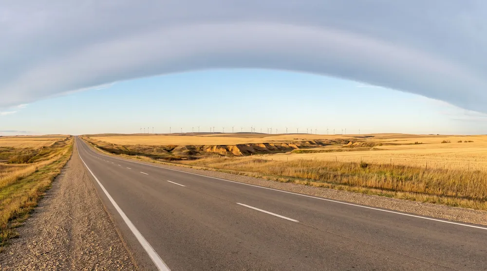 A straight prairie highway in southern Alberta under a long arched chinook cloud, coulees and wind turbines on the horizon